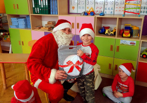 Chłopiec w czerwonej bluzce i czapce Mikołaja pozuje z Mikołajem i prezentem
