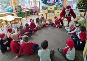 Dzieci ubrane na czerwono siedzą w kole na dywanie i rozmawiają ze Świętym Mikołajem
