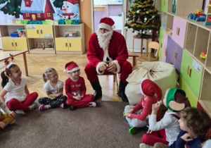 Dzieci ubrane na czerwono siedzą w kole na dywanie i rozmawiają ze Świętym Mikołajem
