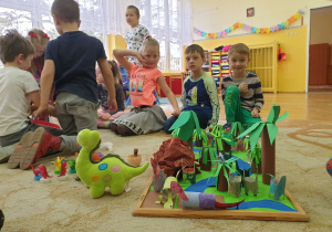 Dzieci siedzą na dywanie w pobliżu makiety z krainą dinozaurów