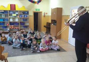 Dzieci słuchają artysty, grającego na puzonie.
