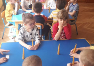 Dzieci siedzące przy stołach, trzymają w rękach świeczkę z wosku pszczelego