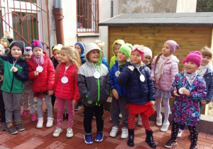 Dzieci z grupy "Mróweczki" stoją na tarasie przedszkolnym. Każde z dzieci ma zawieszony na szyi medal przedszkolaka.