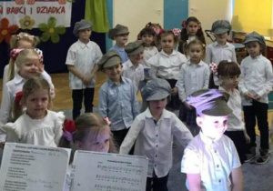 Dzieci z grupy III stoją w rozsypce, ubrane w stroje galowe, chłopcy z czapkami na głowie