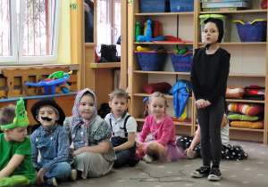 Dziecko przebrane za kotka stoi na "scenie przedszkolnej" obok siedzących dzieci.