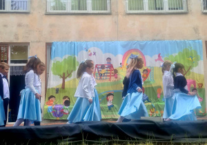 Dzieci na tarasie, dziewczynki ubrane w białe bluzki i niebieskie spódnice, chłopcy w białe koszule i czarne spodnie. Dzieci tańczą walca. W tle dekoracja przedstawiająca plac zabaw.