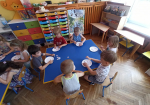 Dzieci siedzą przy stołach i naklejają obrazki na papierowy talerz