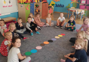 Dzieci siedzą na dywanie. Pośrodku leżą różnokolorowe kropki.