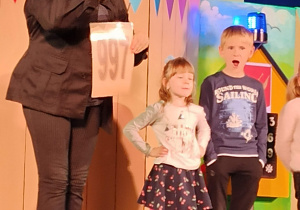 Dzieci na scenie teatru w obecności aktorki, trzymającej kartkę z numerem alarmowym