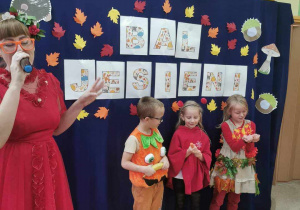 Dzieci przebrane w jesienne stroje stoją na tle dekoracji o tematyce jesiennej, obok stoi prowadząca bal - "Pani Jesień"