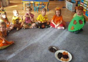 Dzieci przebrane w stroje jesienne siedzą na dywanie.Przed nimi talerz z suszonymi owocami