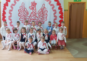 Dzieci ubrane na galowo pozują do zdjęcia na tle dekoracji przedstawiającej Orła Białego