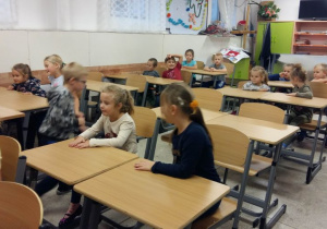 Przedszkolaki siedzą w szkolnych ławkach i słuchają informacji o placówce szkolnej. W tle sali widać meble szkolne.