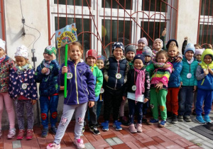 Dzieci z grupy „Leśnych duszków” stoją na tarasie w ogrodzie przedszkolnym. Jedna z dziewczynek trzyma w ręku emblemat umocowany na kijku, przedstawiający napis z nazwą grupy. Reszta dzieci stoi na tle okien jednej z sal przedszkolnych.
