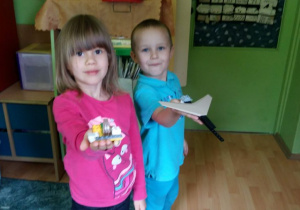 Dziewczynka ubrana w różową bluzkę trzyma w ręku budowlę wykonana z klocków LEGO. Chłopiec ubrany w błękitną bluzkę i getry ma w ręku samolot wykonany z klocków LEGO. W tle widoczne są meble przedszkolne.