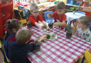 5 dzieci siedzą przy stoliku, nakrytym ceratą w biało-różową kratę. Na stole widoczne są słoiki, wypełnione ziemią i rośliny do posadzenia. W tle widoczny jest kącik z zabawkami i czerwone półki w kąciku przyrody.
