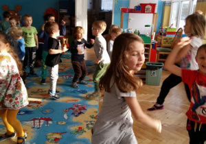 Dzieci z dwóch grup (trzzeciej i piątej) uczestniczą w zabawie andrzejkowej. Tańczą w parach lub rozsypce w sali przedszkolnej. Na zdjęciach widać rozbawione dzieci. W tle widoczne są meble przedszkolne i zabawki.