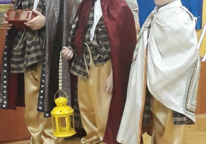 Trzech chłopców ubranych jest w stroje królów. Każdy z chłopców ma na sobie pelerynę, beżowe spodnie. Na głowach mają korony. W rękach trzymają lampion i pudełko.