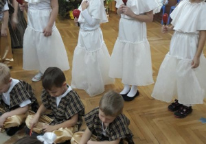 Dzieci siedzą na dywanie w 2 rzędach. Przygotowują się do gry na instrumentach- dzwonkach. Za nimi stoją dziewczynki z marakasami. Dziewczynki przebrane są w białe spódniczki, bluzki i kołnierze a chłopcy mają na sobie ubrane kraciaste kamizelki i beżowe spodnie.