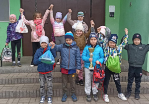 Dzieci stoją przed budynkiem szkoły, trzymając ręce uniesione w górę