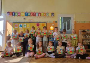Dzieci trzymają pracę plastyczną na tle tablicy z figurami geometrycznymi