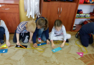 Dzieci siedzą na dywanie w kole. Przed sobą maja rozłożone figury geometryczne i taśmy krawieckie (miarki). Dzieci przykładają taśmy krawieckie do figur, odczytują długość, szerokość figur i zapisują na figurze odpowiednie wyniki pomiarów.