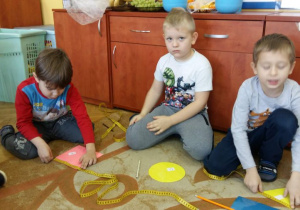 Dzieci siedzą na dywanie w kole. Przed sobą maja rozłożone figury geometryczne i taśmy krawieckie (miarki). Dzieci przykładają taśmy krawieckie do figur, odczytują długość, szerokość figur i zapisują na figurze odpowiednie wyniki pomiarów. Jeden z chłopców, przerywa na moment zadanie.