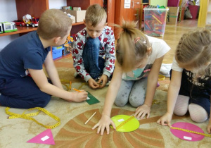 4 dzieci siedzi na dywanie w kole. Przed sobą mają rozłożone figury geometryczne i taśmy krawieckie (miarki). Dzieci przykładają taśmy krawieckie do figur, odczytują długość, szerokość figur i zapisują na figurze odpowiednie wyniki pomiarów.