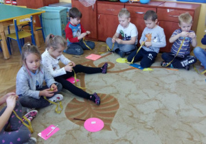 Dzieci siedzą na dywanie w kole. Przed sobą mają rozłożone figury geometryczne i taśmy krawieckie (miarki). Dzieci zwijają miarki krawieckie po skończonej zabawie matematycznej, polegającej na odczytywaniu długośći, szerokości figur.