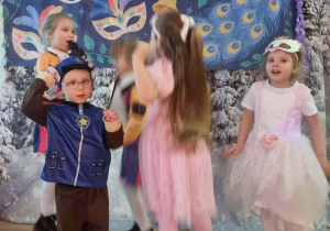 Dzieci przebrane w karnawałowe stroje tańczą na tle dekoracji z napisem Bal Karnawałowy
