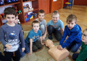 Chłopcy siedzą na dywanie. Jeden z chłopców kładzie ręce na fantomie reanimacyjnym, przygotowując się do uciśnięć klatki piersiowej. Reszta dzieci patrzy na ratownika medycznego.