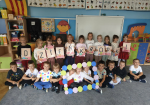 Dzieci pozują do zdjęcia w sali przedszkolnej. Część z nich trzyma w rękach ramkę ozdobioną kropkami a część litery tworzące napis: dzień kropki