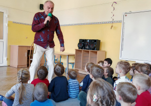 Pan Witek stoi z mikrofonem zwracając się do siedzących przed nim dzieci