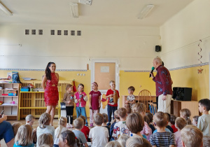 Piątka dzieci stoi pomiędzy tancerką w czerwonej sukience a Panem Witkiem. Trzymają w rękach kubańskie instrumenty. Pan Witek trzyma mikrofon, zwracając się do dzieci.